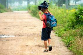 child boy with bag over head walks on soil road at summer