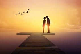 Black silhouettes of the kissing couple on the pier, and flying birds at beautiful and colorful sunset background