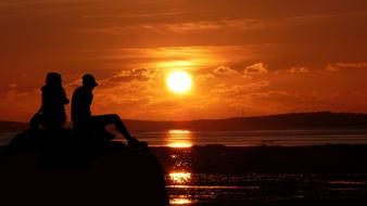 man and woman sitting on stone in front of sea at sunset