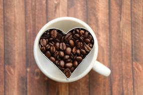 Beautiful cup of coffee with the heart, made of the brown coffee beans, on the wooden surface