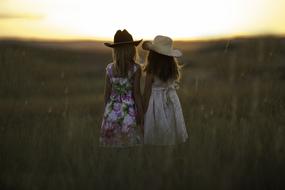 Sisters in colorful dresses and hats, on the beautiful field, at colorful dusk