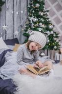 New Yearâs Eve, happy child girl with book sits on bed at christmas tree