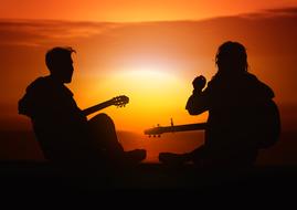 Silhouettes of the couple, playing on guitars, on the shore, at beautiful and colorful sunset on horizon