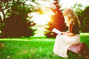 Blonde girl, reading a book on the beautiful and colorful field with trees, at colorful sunset