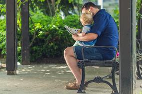 Father with the child reading a book, while sitting on the bench near the green plants