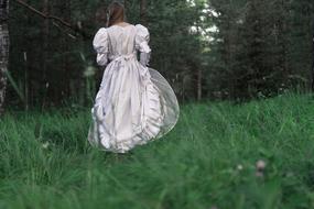 girl in bridal dress walking away on grass to forest