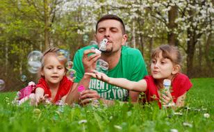 father and two child daughters laying on lawn play with Soap bubbles