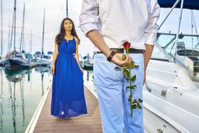 man with rose behind back waiting on beautiful girl at yacht