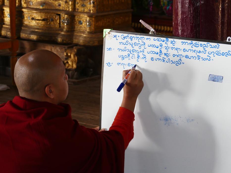 Monk in red robe, writing blue text, on the white board, in Burma