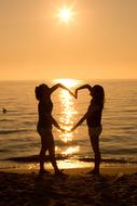 Silhouette of two girls on beach holding hands in Heart shape at Sunset