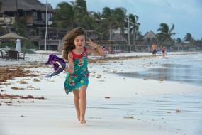 Kid girl, running on the sandy beach, with the people, near the houses and palm trees