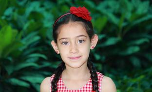 Portrait of a smiling, cute little girl with beautiful, red flower on the head, among the green plants