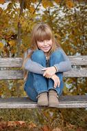 Smiling girl child, sitting on the wooden bench, near the colorful plants in autumn