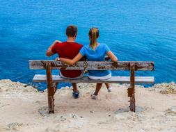 Back view of the couple, sitting on the wooden bench, near the beautiful, blue water