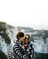 Smiling girls, with the cup of drink, covered in plaid, on the beautiful mountain with green plants
