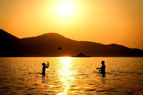 Silhouettes of the people playing in the water, at colorful and beautiful sunset, near the mountains