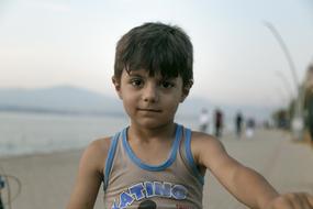 Portrait of the cute child boy on the bicycle, on the beach near the other people, at background with the mountains