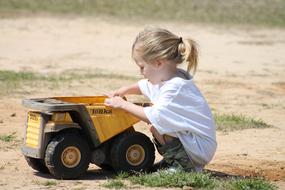 Cute, blonde child girl playing with the yellow and black toy truck, among the green grass outdoors