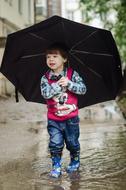 Boy with umbrella, walking on the puddle, near the buildings and plants