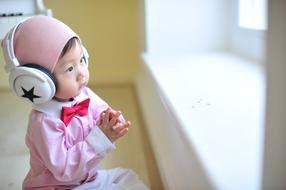 Cute baby in pink hat, with red bow tie and headphones, near the window with light