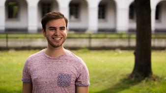 Portrait of the smiling guy with the beard, on the colorful grass with the tree, in sunlight, near the building