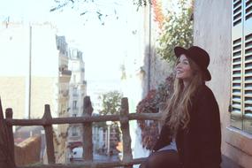 Beautiful, smiling, blonde girl in hat, sitting near the building with the fence