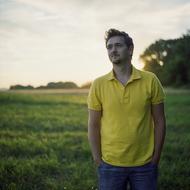 Standing guy with beard, in yellow shirt, on the field with green grass, near the trees, at colorful and beautiful sunset