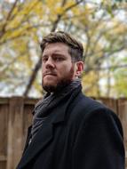 Portrait of the bearded man in coat, near the wooden wall, among the colorful and beautiful trees in the autumn