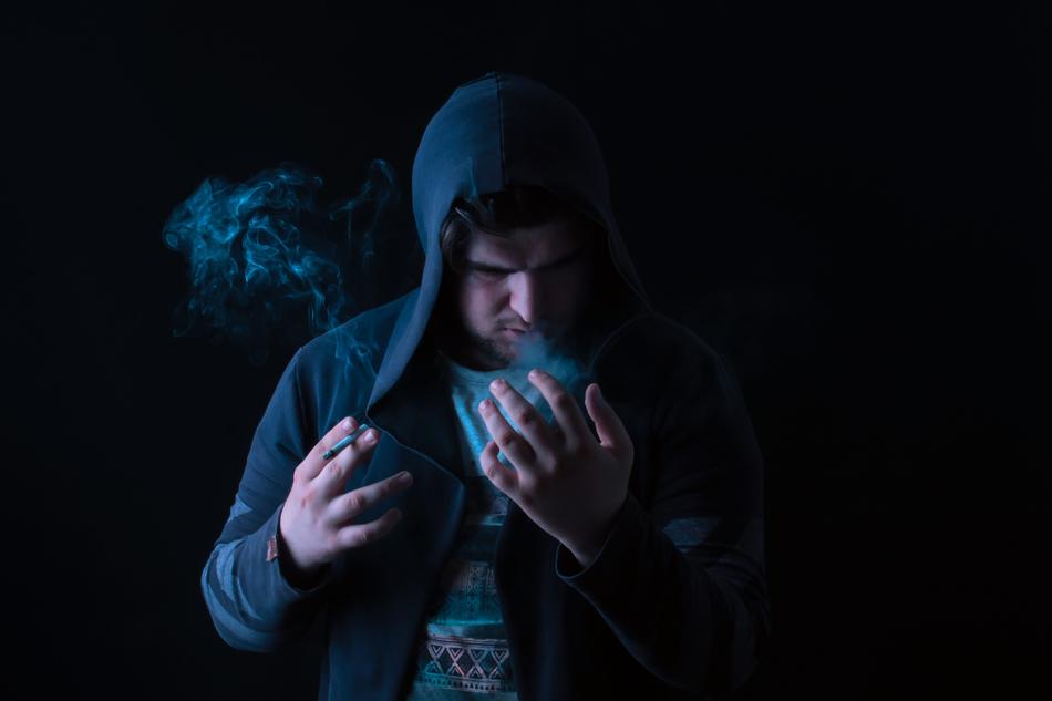 Man, covered in hood, with the smoke on the hands, at black background