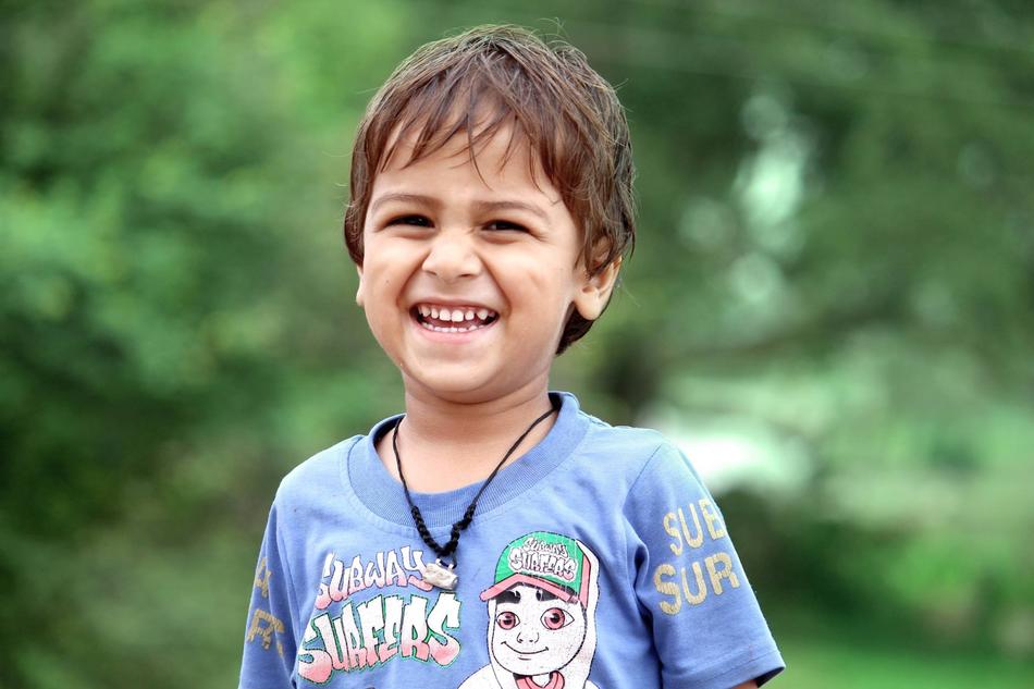 Portrait of the cute, laughing boy with necklace, among the green plants, at blurred background