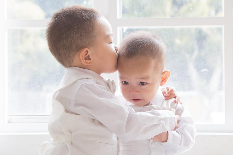 Child brother, kissing another child brother, near the windows with view of the plants and light