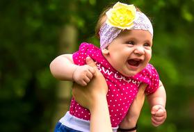 Cute girl in colorful clothing, with yellow flower on the head, in hands of the parent, near the plants