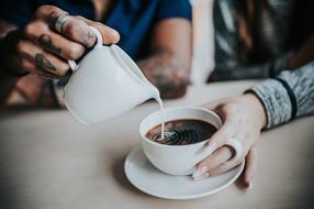 male hand with tattoo pouring Milk to cup with coffee in young female hand