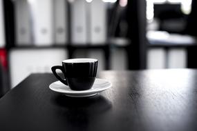 Beautiful, shiny black coffee cup, on the white saucer, on the wooden table, in light and shadow