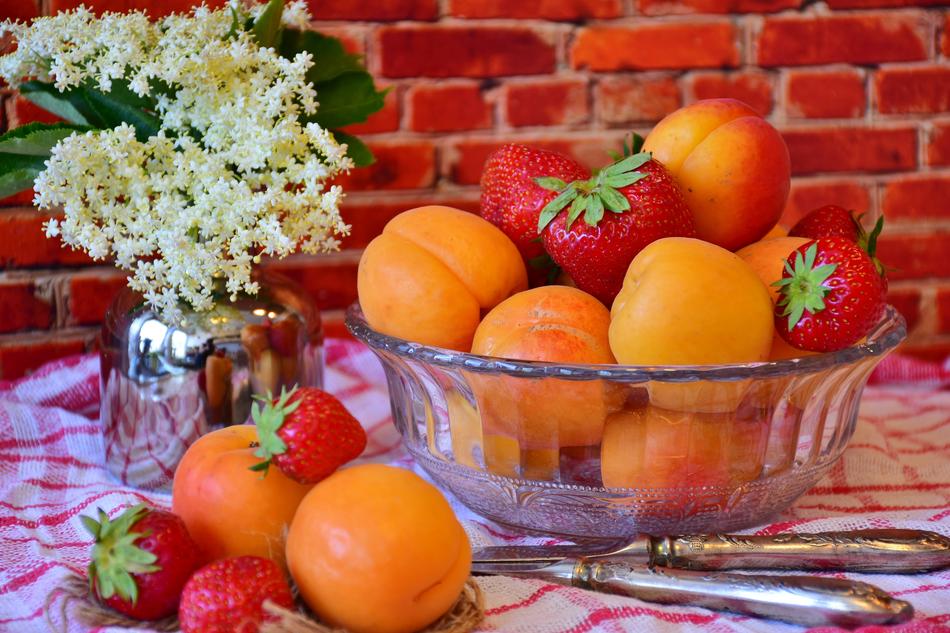 Beautiful and colorful strawberries and apricots in the glass bowl, near the white flowers, near the brick wall