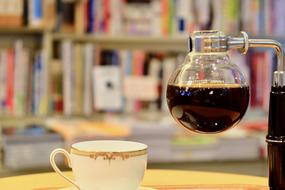 Beautiful, white and gold coffee cup with the pattern, and coffee in the shiny, glass siphon, near the colorful books