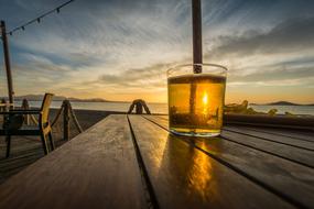 Glass of beer on the wooden table, on the beach, at colorful and beautiful sunset with clouds