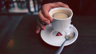 Person holding cup of coffee above the saucer with shiny spoon, on the table