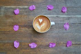 Top view of the cup of coffee, with the beautiful drawing, among the beautiful, purple flower petals, on the wooden surface