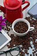 White cup with beautiful, brown coffee beans, near the red pitcher, among the other stuff