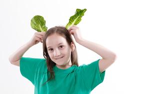 Smiling girl showing rabbit ears with lettuce, at white background