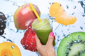 Person, holding green smoothie in cup, at background with colorful and beautiful fruits in water