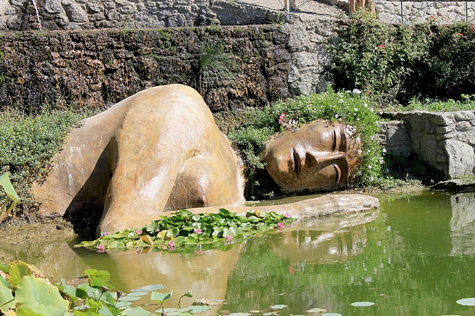 Statue of the laying person, near the water, among the plants and rocks