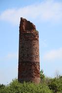 Old, brick chimney of a fireplace, among the green plants, at blue sky with white clouds on background in East Frisia, Germany