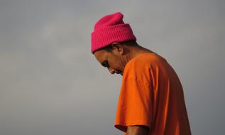 Profile portrait of the man, in orange t-shirt and red beanie, at cloudy sky on background