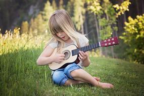 Blonde girl child playing on the guitar on the green grass among the colorful plants on beautiful landscape