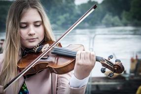 Blonde girl, playing on the beautiful, brown, wooden violin, near the water, among the green trees