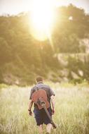 man with guitar on his back walks through Grass in Countryside