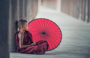 young Buddhist Monk sits on ground near red umbrella