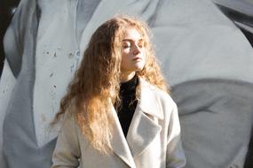 Beautiful, posing girl with curly hair, with closed eyes, wearing white coat, in light
